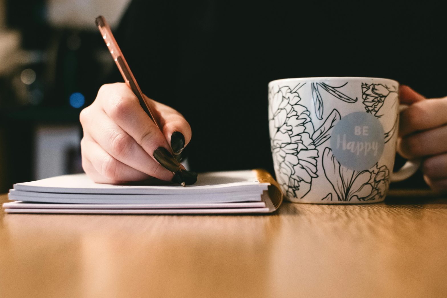 A person writes in a notebook with a pen while holding a white mug with floral designs and the words "Be Happy.