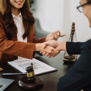 Two people in business attire shake hands across a desk with a contract, gavel, and scales of justice visible.
