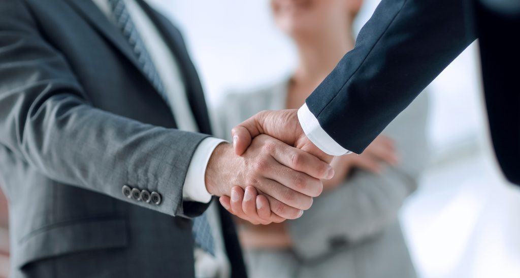 Two people in business suits shake hands, with a third person in professional attire standing in the background.