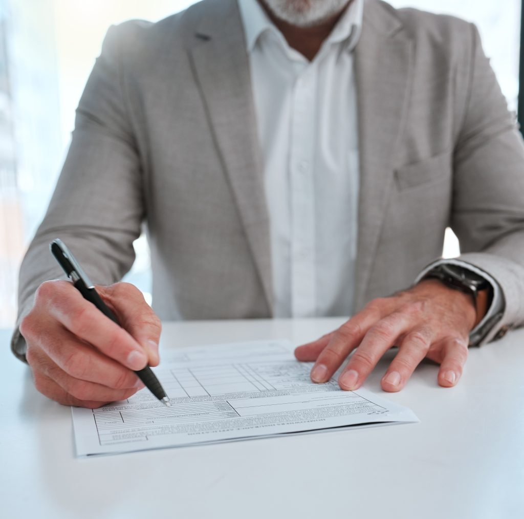 A man in a light gray suit is seated at a desk, filling out a form or document with a black pen.