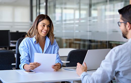A woman holding papers smiles while sitting across from a man with a laptop in a modern office setting.